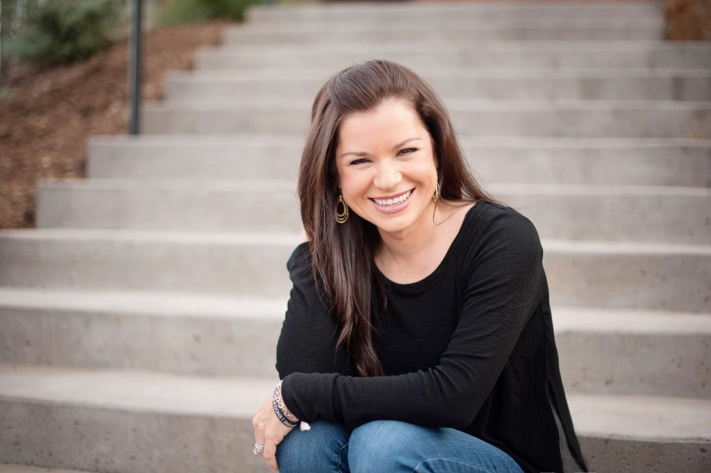 Kaycie LaGrone the interior designer, sitting on concrete steps smiling with her long, brown hair falling down past her shoulders.